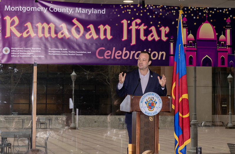 El presidente del Concejo Andrew Friedson habla en el podium en frente de una pancarta que dice “Celebración de Ramadan Iftar en el Condado de Montgomery’ o en inglés “Montgomery County Ramadan Iftar Celebration”.