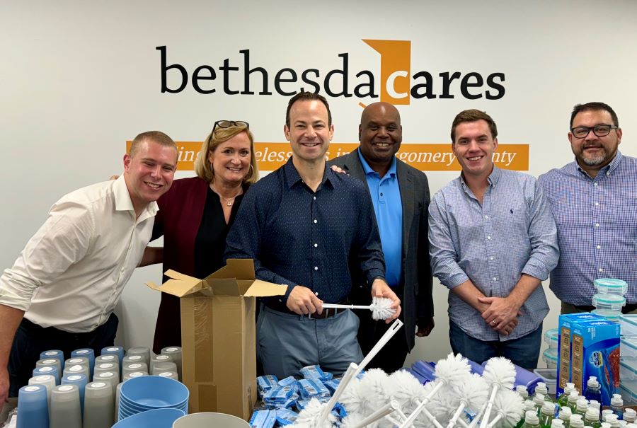 Foto en grupo del equipo de Andrew Friedson y el equipo de la organización conocida en inglés como Bethesda Cares parados en la mesa con artículos para el hogar.