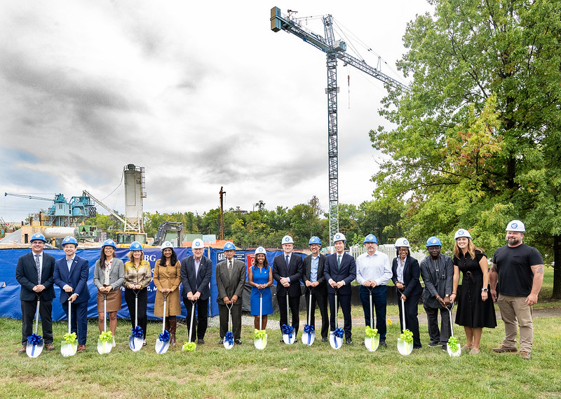 Groupe portant des casques de chantier et tenant des pelles ornées de rubans devant un chantier de construction lors d'une cérémonie de lancement des travaux.