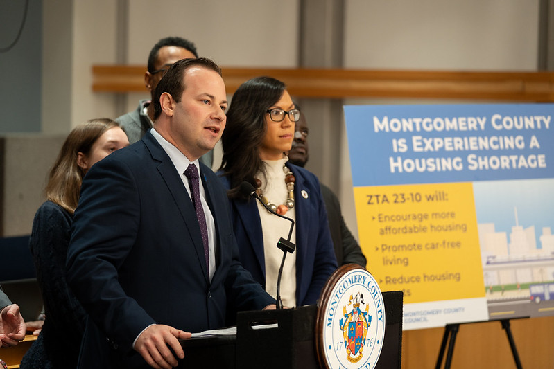 El presidente del Concejo Andrew Friedson habla en un podium al lado de un cartel que traducido al español dice “El Condado de Montgomery está experimentando una escasez de vivienda”