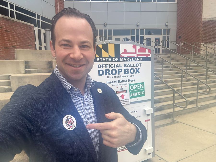 Andrew Friedson usando una calcomanía que dice traducido al español “yo vote” en inglés “I voted” en frente de una urna de votación.