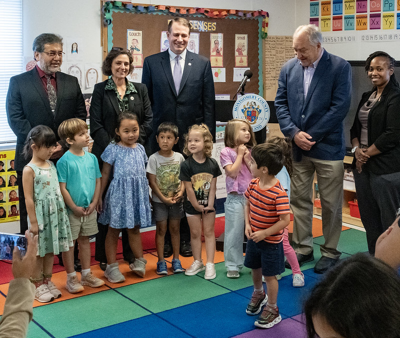 Oficiales de MCPS y del Condado se paran frente de un salón de clases con 8 estudiantes de educación temprana.