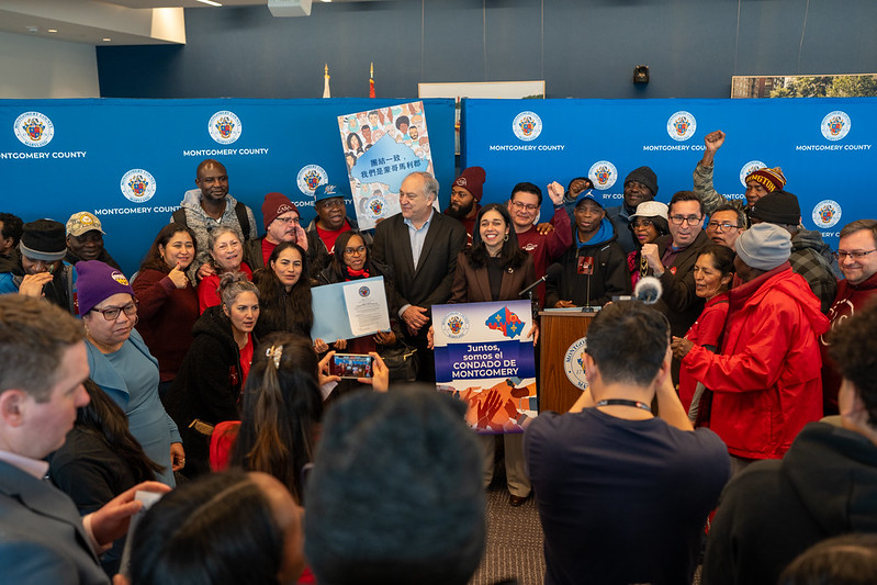 Council President Natali Fani-González, CE Elrich and community members hold up signs at The Trust Act bill signing ceremony. Sign in Spanish reads: “Juntos, somos el Condado de Montgomery”