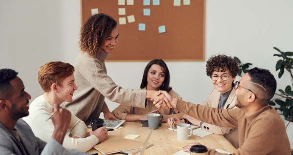 Two people shaking hands across a table, with other people seated at the table looking on.