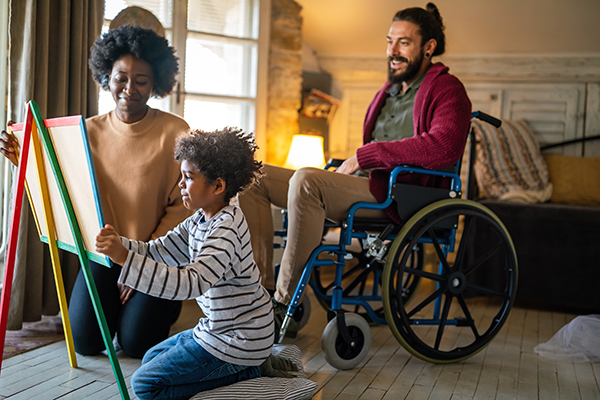 a dad who is using a wheelchair is smiling at his wife and young son who are making art on a chalkboard in their home