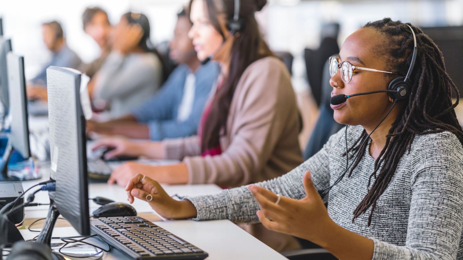 Call center operators at their computers wearing headsets out of focus. The operator in focus is wearing glasses and speaking with a caller.