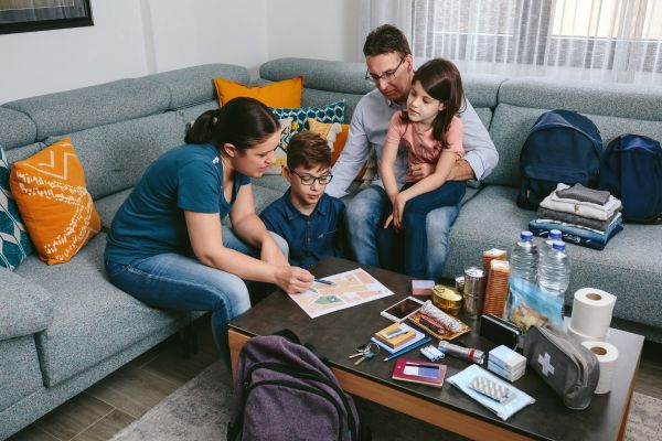 Family in living room making an emergency plan. There are emergency supplies on their living room table: water, food, toilet paper, batteries, flashlight, backpacks, clothing and more.