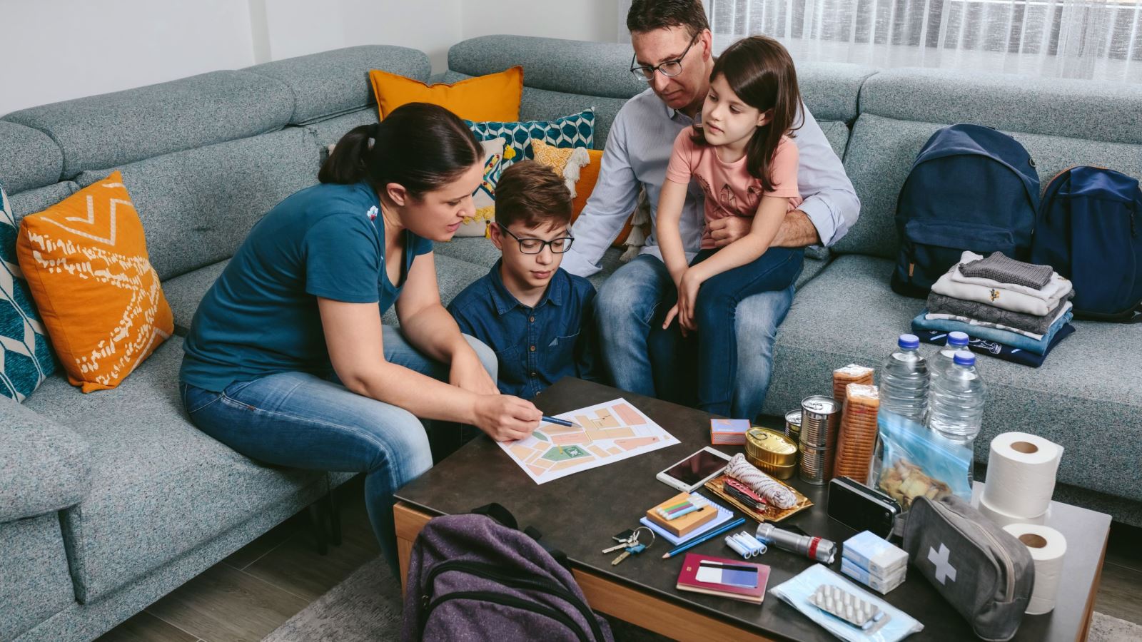 Family in living room putting together and emergency kit with backpacks, clothing, water, toilet paper, food, batteries, identification, flash light, medication, etc.